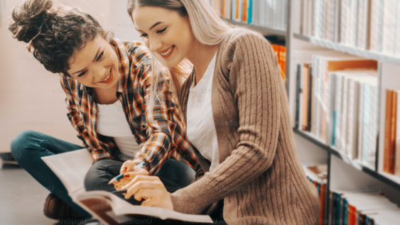 Zwei junge Frauen sitzen in einer Bibliothek auf dem Boden und unterhalten sich über etwas, das in dem Buch steht, das eine von ihnen im Schoß geöffnet hat.