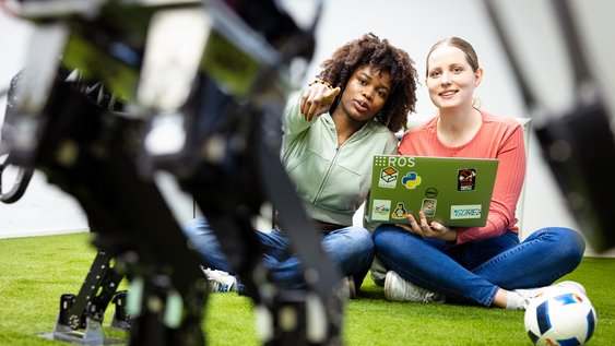 Zwei Studierende sitzen auf dem Boden und schauen auf Roboterfüße