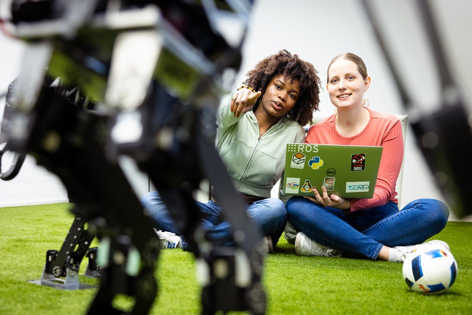 Zwei Studierende sitzen auf dem Boden und schauen auf Roboterfüße